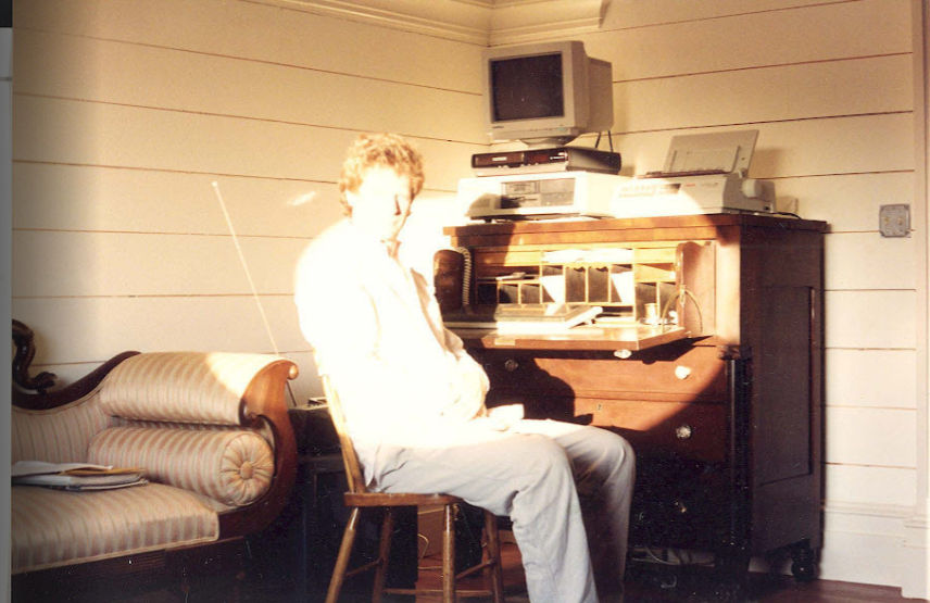 Carter at secretary desk in attic apartment, Windows 95 computer stacked on top, lit by oval window